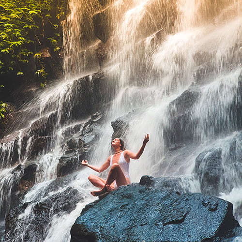 A lady is sitting under the waterfall and meditate with the sound of the water